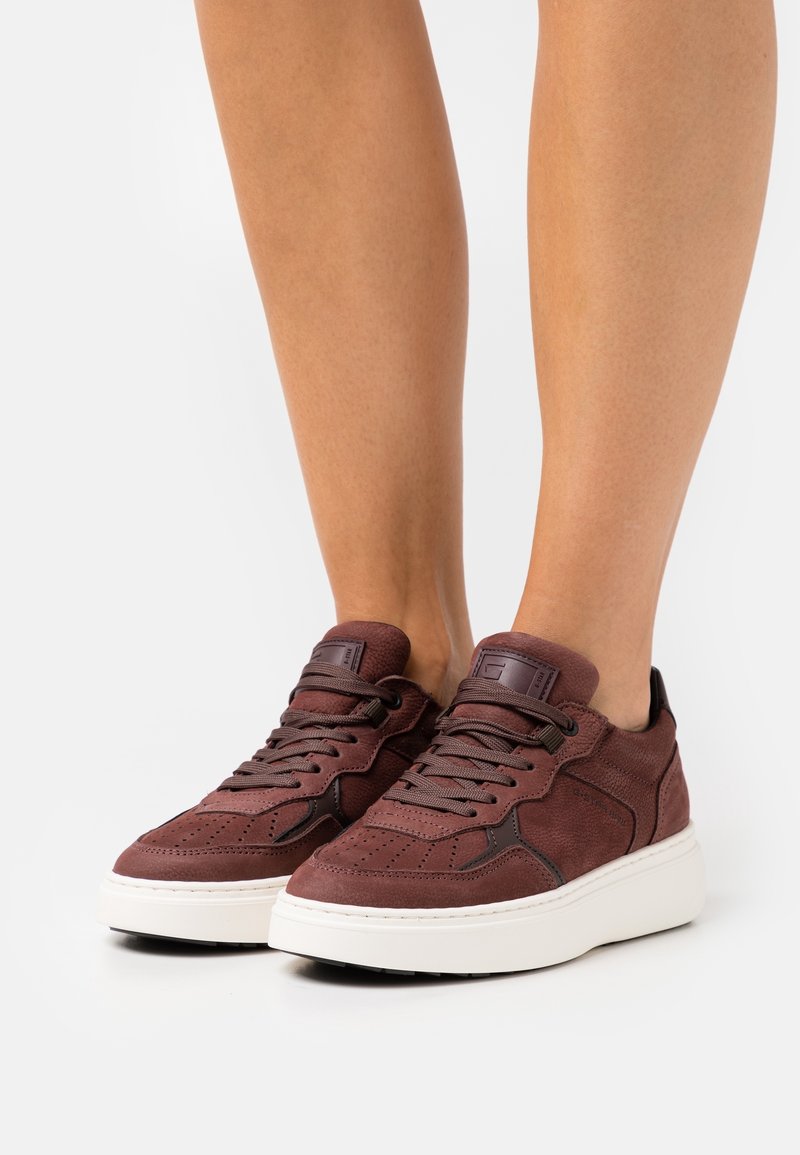 Personne portant des baskets en daim marron à lacets avec des semelles épaisses blanches sur un fond clair uni.