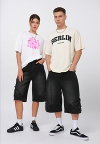 Dos modelos llevan camisetas de gran tamaño y pantalones cortos de mezclilla negros a la altura de la rodilla con detalles desgastados. Un par presenta un gráfico blanco, el otro dice "BERLÍN".