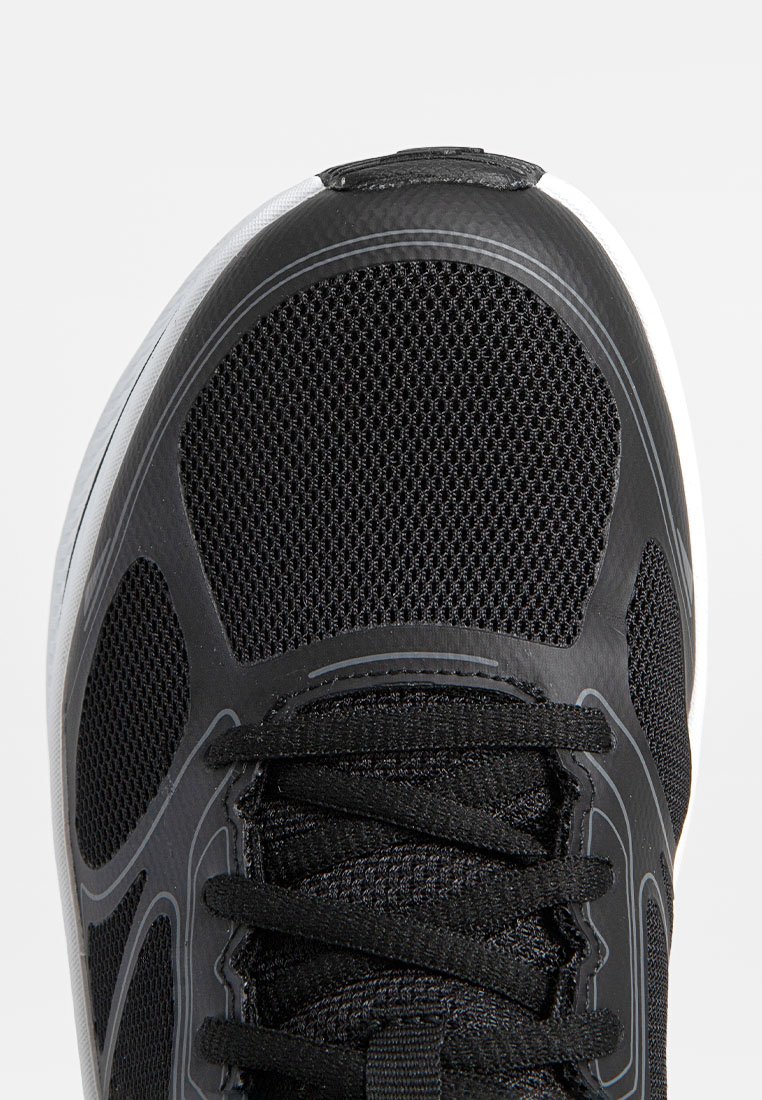 Zapatilla deportiva negra con parte superior de malla, superficie texturizada, puntera redonda y suela blanca contrastante. Presenta cordones negros y acentos de diseño reforzados.