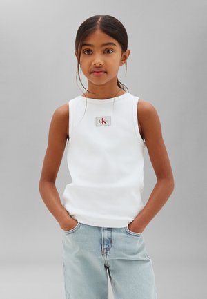 Ung pige med mørkt hår i en hvid ærmeløs top med Calvin Klein-logo, hænderne i lommerne på lyseblå jeans, neutral grå baggrund.