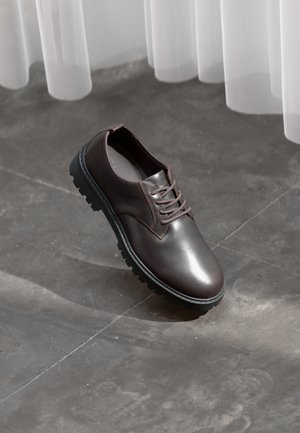 Brown leather lace-up shoe with thick black sole on a concrete floor beneath sheer white curtains.