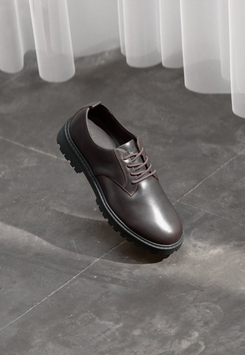 Brown leather lace-up shoe with thick black sole on a concrete floor beneath sheer white curtains.
