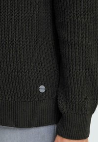 Pulover tricotat de culoare verde închis, cu cusături texturate, manșete și tiv cu dantelă, având un eticheta rotundă cu logo aproape de margine.