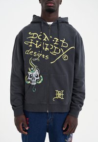 Felpa con cappuccio grigia con cerniera, con testo grafico giallo, teschio e disegni di serpente. Tessuto in misto cotone con tasche frontali e cappuccio regolabile.