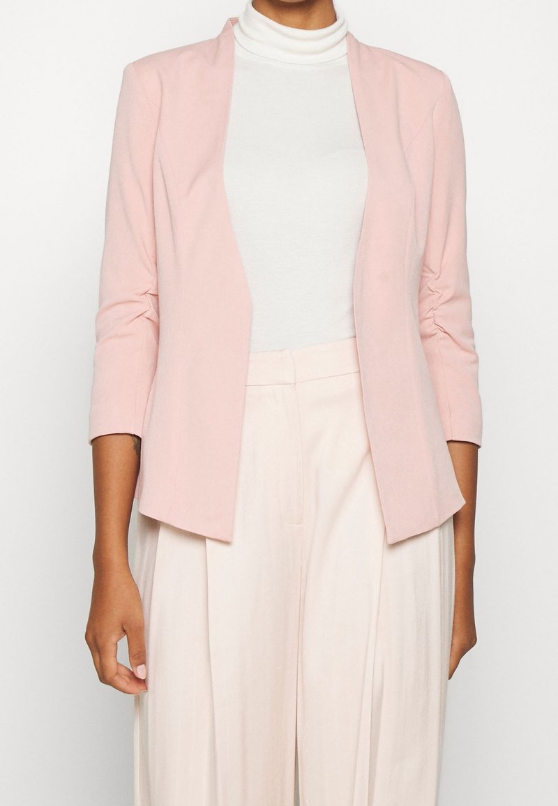 Blazer rose clair à coupe décontractée, ouvert sur le devant, avec des manches trois-quarts, associé à un col roulé blanc et un pantalon clair.