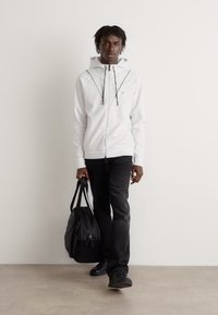 Haut à glissière blanc avec des cordons de serrage noirs, associé à un jean noir et des chaussures noires. Portant un sac fourre-tout noir, se tenant contre un fond uni.