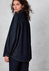 Göndör hajú nő sötét kék farmerovershirtben és hozzá illő nadrágban, vállát hátrafordítva néz egy sima háttér előtt.