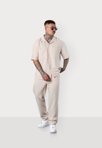 Short-sleeve, collared, light beige patterned shirt paired with matching tapered trousers and white sneakers. Smooth textile, relaxed fit.