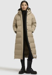 Beige pufferjas met een lange snit, oversized capuchon, zwarte voering, drukknoopsluiting en zijzakken; gecombineerd met zwarte laarzen.