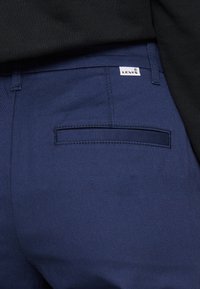 Pantalons bleu marine en tissu texturé, avec une poche arrière et une étiquette logo Levi's sur la ceinture.