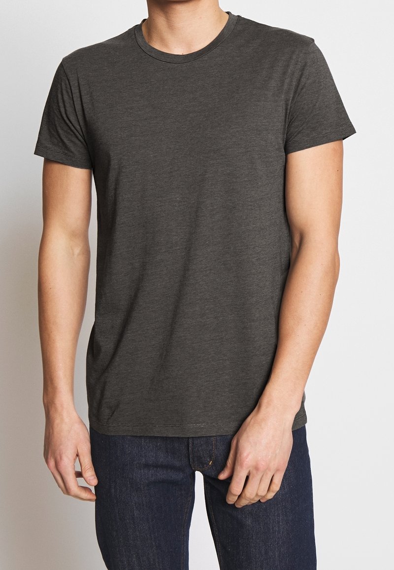 Homme portant un t-shirt gris foncé uni à manches courtes et un jean bleu foncé, debout devant un fond neutre.