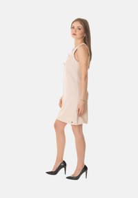 Abito beige senza maniche con un dettaglio a fiocco sul collo, realizzato in tessuto morbido, abbinato a scarpe con tacco alto nere, design minimalista.