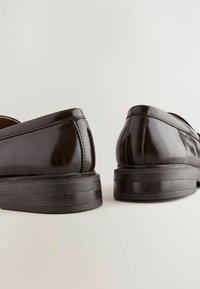 Dunkelbraune, glänzende Slipper mit glatter Textur, genähten Details und einer robusten schwarzen Sohle, die ein klassisches, rundes Design unterstreichen.