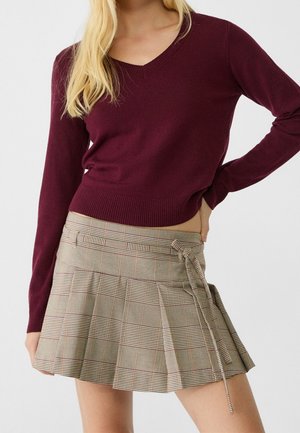 Femme portant un pull bordeaux à col en V et manches longues ainsi qu'une mini-jupe plissée beige à carreaux avec un lien sur le côté.