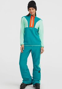 Teal Ski-Pullover mit hohem Kragen, ausgestattet mit einem orangefarbenen Reißverschluss und hellen Minz-Akzenten; kombiniert mit teal-farbenen Schneehosen für den Wintersport.