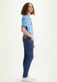 Camisa de manga corta azul con estampado, vaqueros oscuros, zapatillas blancas. El modelo está de perfil, mostrando el conjunto y los detalles, incluido un parche con logo.