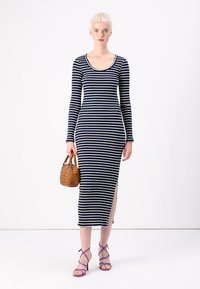 Μακριά φόρεμα με ριγέ navy και λευκό σχέδιο, μακριά μανίκια και πλάγιες σχισμές; μοντέλο που κρατά καφέ τσάντα, φορά μωβ σανδάλια με λουριά. Λείο, εφαρμοστό ύφασμα.