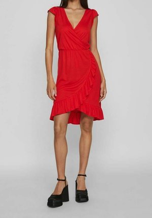 Kobieta w czerwonej sukience kopertowej do kolan z falbaną i czarnych platformowych szpilkach, stojąca na tle jednolitego szarego tła.