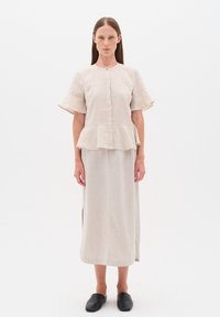 Béžový lněný peplum top s krátkými, rozšířenými rukávy a zapínáním na knoflíky, spárovaný s odpovídající dlouhou sukní s bočními rozparky. Černé ploché boty.