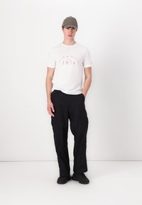 Tricou alb cu mânecă scurtă și imprimeu roșu, pantaloni cargo negri, șapcă gri cu text și adidași negri. Design simplu cu un stil funcțional.