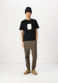 Sort kortærmet t-shirt med et mønstret grafik på forsiden, parret med brune tapered bukser og brune sneakers. Sort strikket beanie tilbehør.