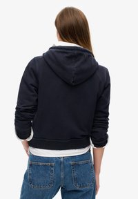 Marineblå cropped hoodie med en snøre i hætten, manchetter og ribbede kanter. Båret over en hvid skjorte med krave, parret med højstående denimjeans.
