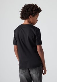 Juodas medvilninis marškinėliai trumpomis rankovėmis, apvaliu kaklu ir paprastu dizainu, dėvimi su pilkais džinsais. Žiūrima iš nugaros.