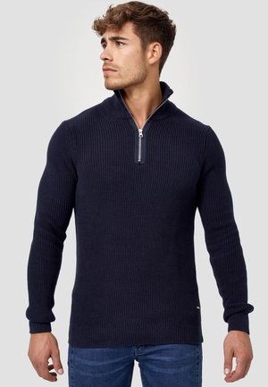 Marineblauer Reißverschluss-Pullover mit gerippter Textur, langen Ärmeln, hohem Kragen und silbernem Reißverschluss-Akzent. Getragen mit blauen Jeans.