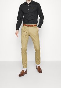 Uomo che indossa una camicia nera a bottone, pantaloni beige con cintura marrone, calze bianche e mocassini marroni, in piedi contro uno sfondo bianco semplice.