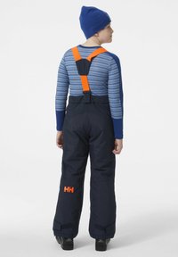 Mono de invierno azul marino con tirantes naranjas ajustables, rodillas reforzadas y puños de Velcro. Camiseta de manga larga azul marino con rayas azules debajo.