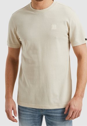 Beige tekstureret kortærmet t-shirt med lille firkantet logo på venstre bryst båret af en person i blå jeans.
