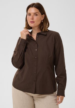 Femme aux cheveux bruns mi-longs porte une chemise marron boutonnée et un pantalon beige, debout devant un fond clair uni.