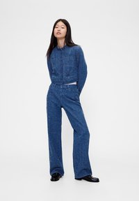 Komplet iz jeans jakne in širokih hlač, modra tkanina z vzorcem, skrajšan kroj, veliki sprednji žepi, kombinirano s črnimi čevlji.