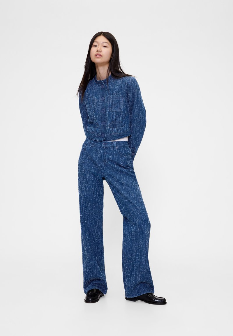 Komplet iz jeans jakne in širokih hlač, modra tkanina z vzorcem, skrajšan kroj, veliki sprednji žepi, kombinirano s črnimi čevlji.