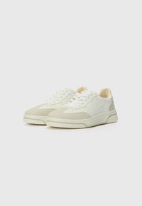 White and light beige sneakers with a textured upper, flat laces, perforated toe, and a thick rubber sole. Minimalist design elements.