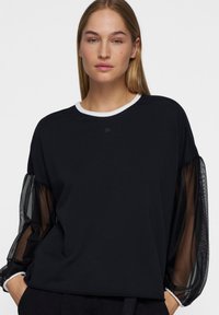 Zwarte shirt met doorzichtige mesh ballonsleeves, ronde witte kraag, zachte stof, losse pasvorm, minimale merkvermelding op de borst.