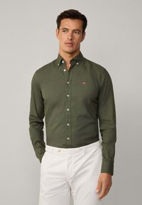Camicia verde oliva a maniche lunghe con bottoni bianchi e un piccolo logo rosso sul petto, abbinata a pantaloni bianchi.