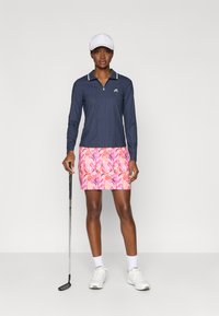 Tumesinine pika varrukaga tõmblukuga jakk valgete aktsentidega, koos roosa lillemustriga seeliku ja golfikepiga. Mall kannab valget mütsi ja kingi.