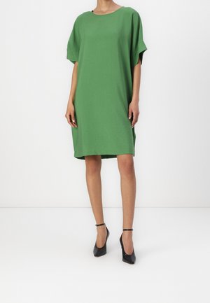 Vrouw draagt losse groene jurk tot op de knie met korte mouwen en zwarte enkellaarsjes met bandje, staand tegen een witte achtergrond.