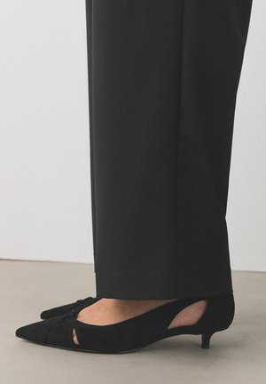 Feet wearing black suede low kitten-heel slingback shoes with cutout details, paired with long black pants on a light floor.