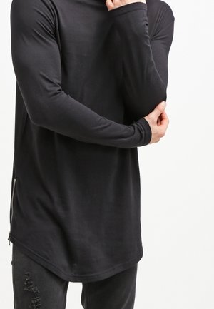 Haut noir à manches longues avec un col montant, une coupe ajustée et un détail de fermeture éclair latérale. Le tissu semble lisse avec une texture légère.