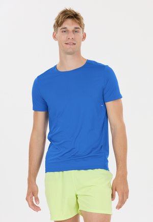 Jeune homme portant une chemise à manches courtes bleu vif et un short vert clair, debout devant un fond blanc uni.