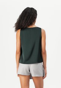 Mujer con cabello largo y rizado que lleva una blusa sin mangas de color verde oscuro y pantalones cortos grises claros, de pie de espaldas contra un fondo blanco.