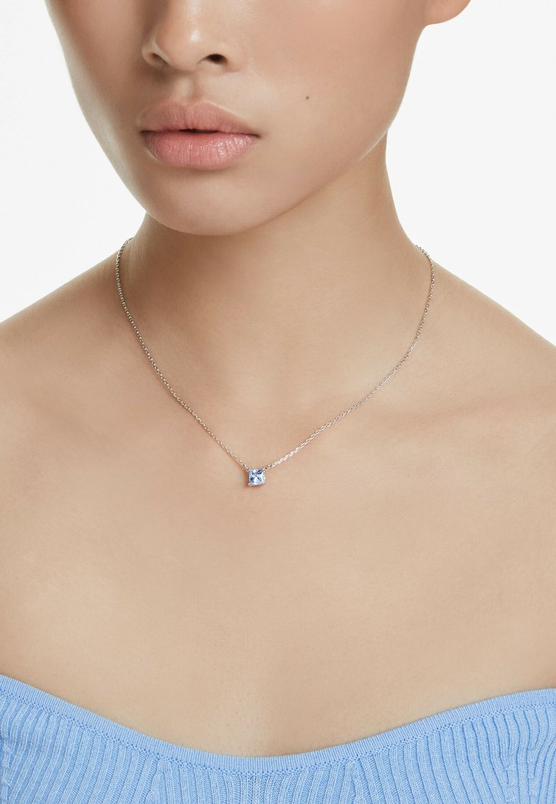 Collana in argento con pendente di pietra blu quadrata. Texture liscia, design delicato, indossata sopra un top a coste blu chiaro.