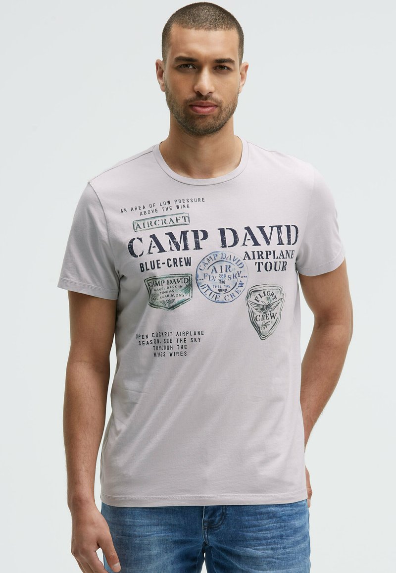 Gray short-sleeve t-shirt featuring printed patches and text related to aviation, with a round neckline and a relaxed fit.