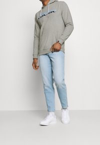 Sudadera gris con logo azul, cordones blancos, bolsillo frontal, combinada con jeans azul claro y zapatillas blancas.