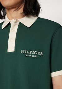 Zelená polokošile s krémovým žebrovaným límečkem, třemi knoflíky a vyšitým nápisem "HILFIGER NEW YORK" bílou barvou na levé části hrudi.
