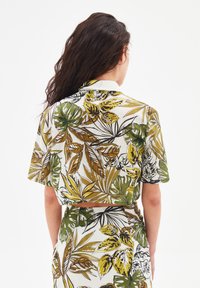 Camicetta accorciata con una vivace stampa botanica multicolore, caratterizzata da grandi foglie verdi e gialle su uno sfondo chiaro. Design con colletto.