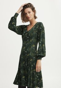 Grünes Kleid mit V-Ausschnitt, puffärmeligen langen Ärmeln und ausgestelltem Saum. Es hat ein Blumenmuster in Gelb und Weiß auf einem dunkelgrünen Hintergrund.