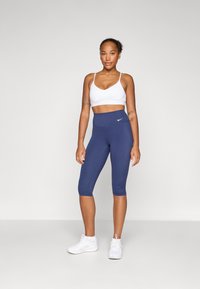 Vit sport-bh och marinblå capri leggings med hög midja, som har en liten vit Nike-logga. Modellen bär vita sportskor.